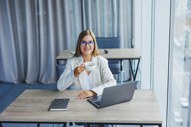 Fröhliche Managerin mit Brille und Laptop lächelt während der Kaffeepause im Büroinnenraum in die Kamera Fröhliche Europäerin mit Koffeingetränk posiert