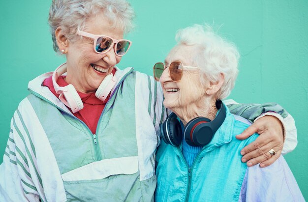 Fröhliche Mode und ältere Freunde umarmen sich in coolen Sonnenbrillen, Vintage-Jacken und modernen Kopfhörern im Freien Retro-Wandhintergrund und ältere Frauen, die sich mit Stolz Freiheit umarmen und im Ruhestand lächeln