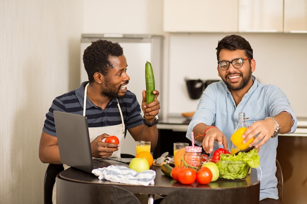 Fröhliche multiethnische Studenten, die gesundes Essen zubereiten und Spaß in der Küche haben. Afroamerikaner und sein indischer Freund sitzen am Tisch und schauen sich eine Kochshow an und lachen. Kochen und Technik.