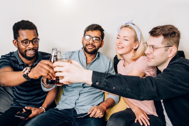 Fröhliche multikulturelle Freunde, Studenten, die Freizeitkleidung tragen, trinken, Flaschen und Gläser klirren, Geburtstag feiern. Drei Mann und Frau, die Spaß in der Heimparty haben.