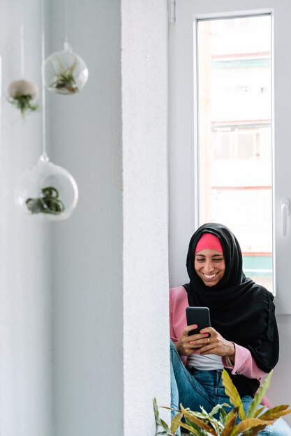 Fröhliche muslimische Frau trägt einen Hijab mit Smartphone drinnen. Vertikale Seitenansicht einer arabischen Frau, die zu Hause Musik mit Kopfhörern neben einem Fenster hört. Technologie und muslimischer Lebensstil