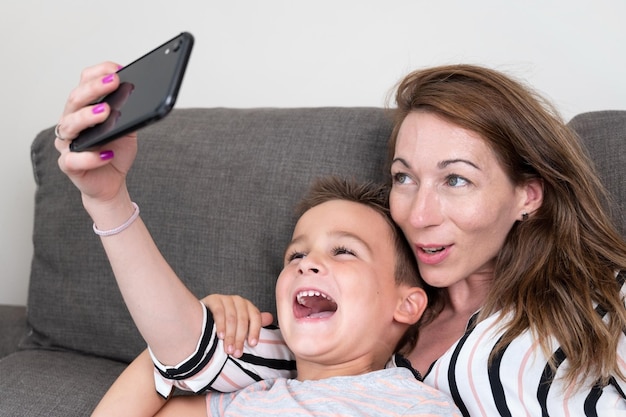 Fröhliche Mutter, die ein Smartphone hält, das Selfie mit ihrem kleinen Sohn macht, der Siegeszeichen zeigt