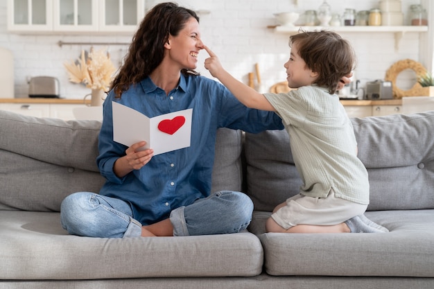 Fröhliche Mutter lachend Spiel mit süßem Sohn halten Geschenkpostkarte zum Geburtstag oder Muttertag auf der Couch