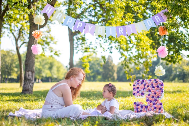Fröhliche Mutter und Tochter, die sich am Kindergeburtstag auf einer Decke mit Papierdekorationen im Park amüsieren