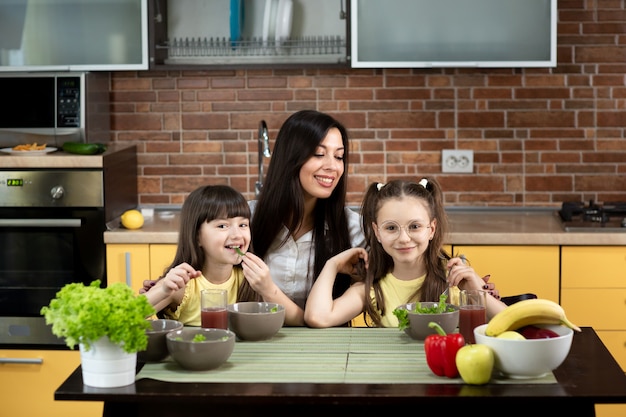 Fröhliche Mutter und zwei Töchter essen zu Hause gemeinsam gesunden Salat. Das Konzept von gesunder Ernährung, Familienwerten, gemeinsamer Zeit