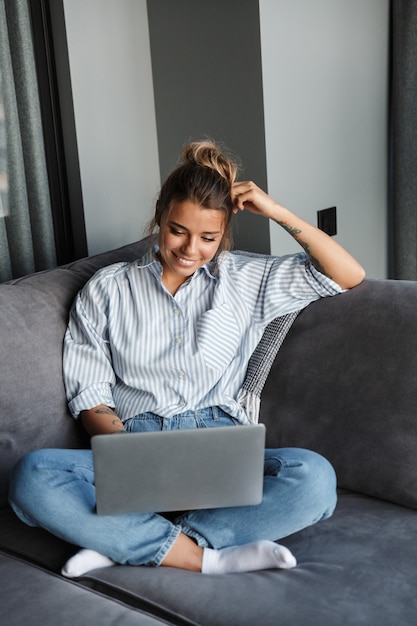 fröhliche nette Frau, die lächelt und Laptop benutzt, während sie auf der Couch im Wohnzimmer sitzt?