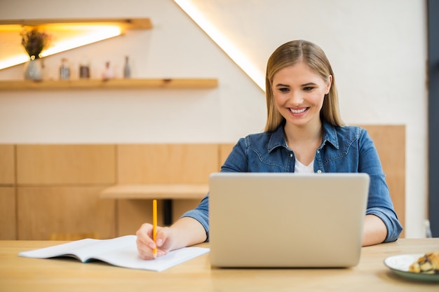 Fröhliche nette kluge Frau, die im Büro sitzt und ihren Laptop während der Arbeit benutzt