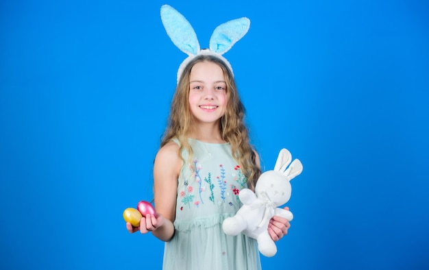 Fröhliche Ostern Fröhliches kleines Mädchen mit Osterhasenohren Kleines Mädchen im Häschenstirnband mit farbigen Eiern und Spielzeug Entzückendes Mädchen mit niedlichem Stoffhasen-Spielzeug Osterhase ist ein Symbol für Ostern
