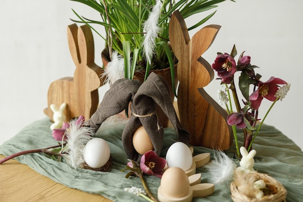Fröhliche Ostern Stilvolle Holzhäschen Frühlingsblumen natürliche Eier und Serviettenhäschenohren auf rustikalem Tisch im Zimmer Osterstillleben Festliche Anordnung und Dekoration im Bauernhaus