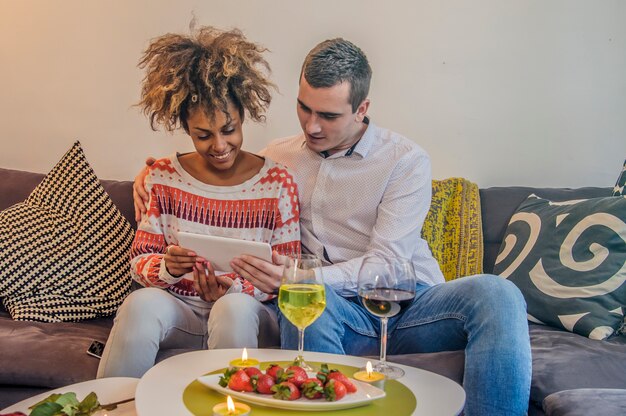 Fröhliche paar Spaß mit Tablette im Sofa. Mixed Paar zu Hause E-Shopping mit Tablette. Mix Race Frau. Happy Couple Sitting Auf Couch Mit Tablet-Computer