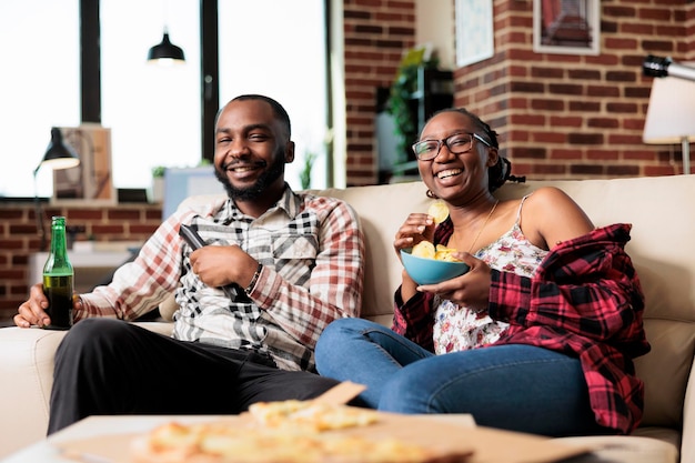 Fröhliche Partner, die Filme im Fernsehen sehen und Essen zum Mitnehmen von der Lieferung essen, zusammen Spaß haben. Fast Food zum Mitnehmen und Bierflaschen genießen, während sie sich einen Film im Fernsehen ansehen.