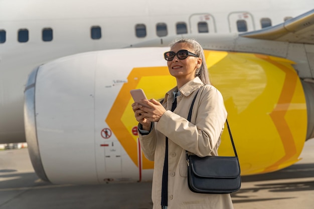 Fröhliche Passagierin mit Sonnenbrille, die draußen am Flughafen in der Nähe des Flugzeugs telefoniert