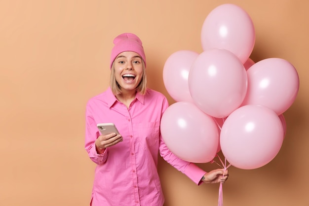 Fröhliche positive Europäerin ruft vor Freude aus, nutzt Handy und bekommt Glückwünsche zu ihrem Geburtstag in rosa Kleidung, hält einen Haufen Luftballons isoliert auf braunem Hintergrund