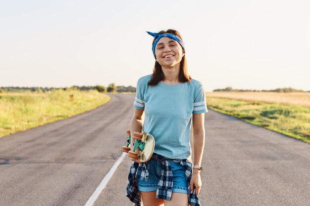 Fröhliche positive Frau, die kurzes, blaues T-Shirt und Haarband trägt, stehend und in die Kamera schaut, Skateboard in den Händen hält und im Sommer Zeit im Freien verbringt.