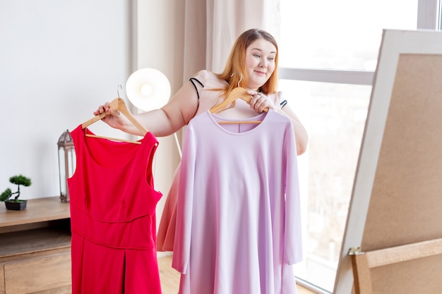 Fröhliche positive Frau, die vor dem Spiegel steht, während sie zwei verschiedene Kleider vergleicht