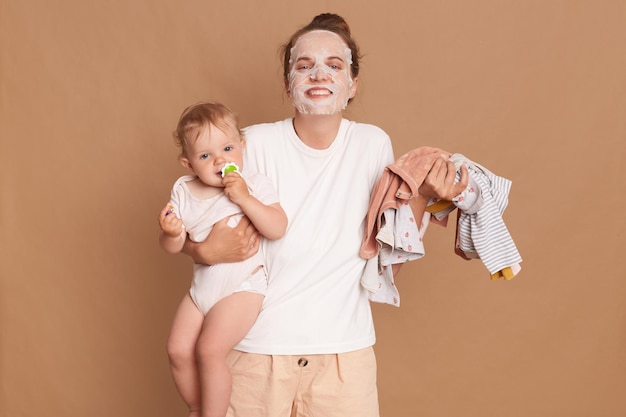 Fröhliche positive Frau mit weißem T-Shirt, die kosmetische Eingriffe durchführt, während sie mit Kindern spielt und Hausarbeiten erledigt, die kleine Tochter in den Händen halten und positive Emotionen ausdrücken