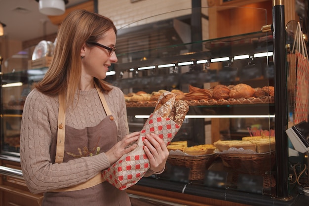 Fröhliche professionelle Bäckerin, die frisches leckeres Brot hält und in ihrer Bäckerei arbeitet