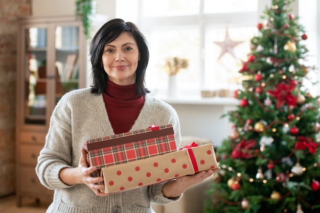 Fröhliche reife brünette Frau in Freizeitkleidung, die einen Stapel Geschenkboxen mit Geschenken für ihre Familie gegen geschmückten Weihnachtsbaum zu Hause hält