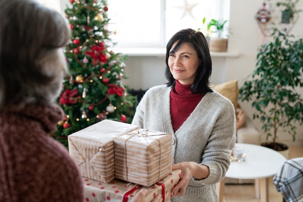 Fröhliche reife Frau, die ihrem Mann verpackte Geschenke gibt, während sie ihn am Fenster in der häuslichen Umgebung gegen den Weihnachtsbaum betrachtet