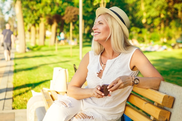 Foto fröhliche reife frau, die sommertag im park sitzt, der auf der bank sitzt