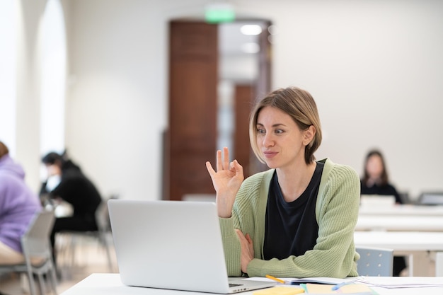 Fröhliche reife Studentin, die während des virtuellen Unterrichts online in der Bibliothek eine Ok-Geste zeigt