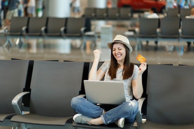 Fröhliche reisende Touristenfrau mit Laptop, die mit gekreuzten Beinen sitzt, Siegergeste hält Kreditkarte wartet in der Lobbyhalle am Flughafen?