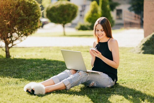 Fröhliche reizende junge Frau, die ein Telefon durchsucht, während auf dem Gras mit Laptop-Computer sitzt, gekleidet in stilvolle Kleidung. Studentenleben. Draußen.