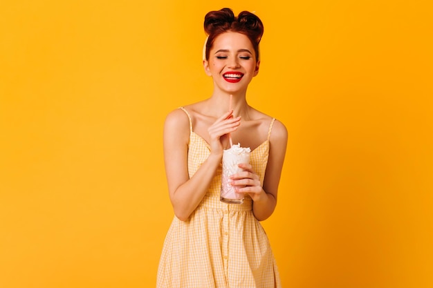 Fröhliche rothaarige Dame im Kleid, die sich auf gelbem Hintergrund amüsiert Kühlendes Pinup-Mädchen, das Milchshake trinkt