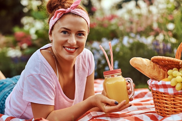 Foto fröhliche rothaarige frau mittleren alters in lässiger kleidung mit einem stirnband, die beim picknick im freien genießt, während sie auf einer decke im park liegt.