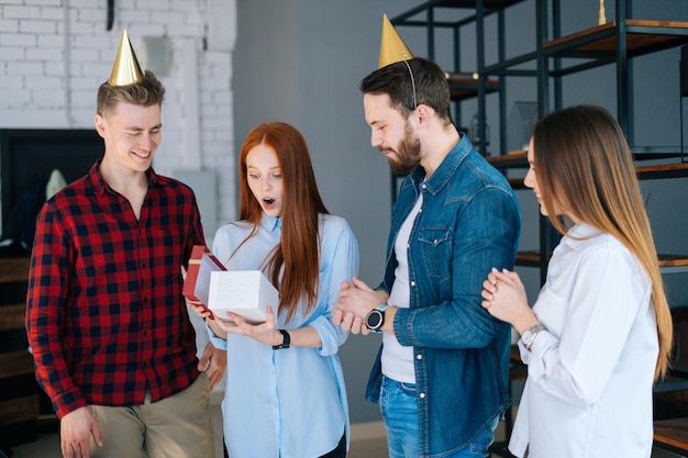 Fröhliche rothaarige junge Frau Eröffnungsgeschenk von Kollegen im Büro zum Applaus der Mitarbeiter Glückliches Team junger Geschäftsleute gratuliert Kollegin und macht Geschenk