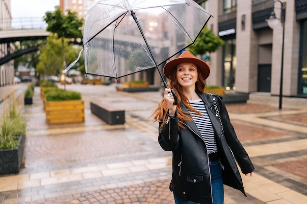 Fröhliche rothaarige junge Frau mit Modehut tanzt und hat Spaß mit transparentem Regenschirm auf einer schönen Stadtstraße und genießt Regenwetter im Freien