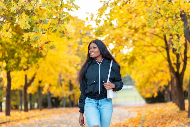Fröhliche schöne afroamerikanische Frau mit einem süßen Lächeln in einer modischen Freizeitjacke mit Blue Jeans und einer Handtasche spaziert in der Natur in der Nähe der Bäume mit leuchtend gelbem Herbstlaub