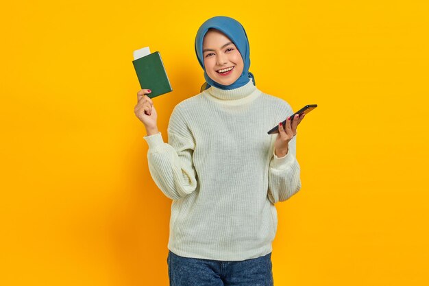 Fröhliche schöne asiatische Frau im weißen Pullover mit Handy und Reisepass isoliert auf gelbem Hintergrund Sommerreise