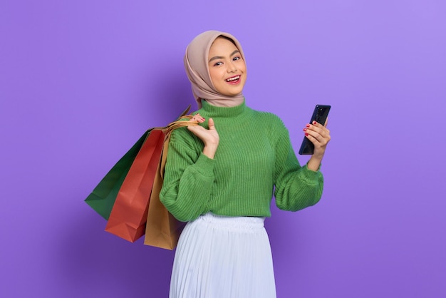 Fröhliche schöne asiatische Frau in grünem Pullover mit Einkaufstüten, mit Handy und Blick auf die Kamera isoliert über lila Hintergrund