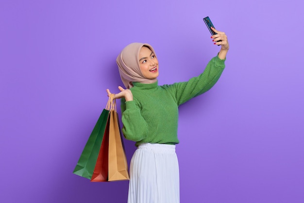 Fröhliche schöne asiatische Frau in grünem Pullover mit Einkaufstüten und macht ein Selfie auf dem Handy einzeln auf violettem Hintergrund