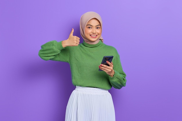 Fröhliche, schöne asiatische Frau in grünem Pullover und Hijab, die ein Mobiltelefon benutzt und den Daumen isoliert über violettem Hintergrund zeigt