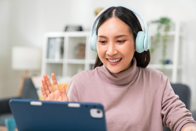 Fröhliche schöne asiatische Frau mit Kopfhörern und Blick auf das digitale Tablet bei Videoanrufen und Online-Studien im Internet.