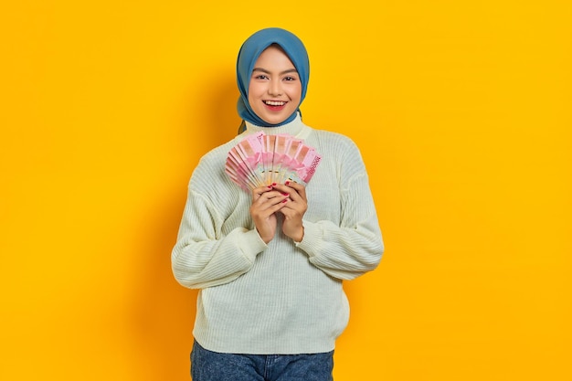 Fröhliche schöne asiatische Muslimin in weißem Pullover mit Geldfächern in Rupiah-Banknoten isoliert auf gelbem Hintergrund Religiöses Lifestyle-Konzept der Menschen
