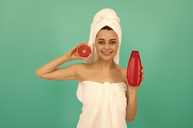 Fröhliche schöne Frau im Handtuch mit Grapefruit-Shampoo-Flasche auf blauem Hintergrund