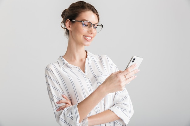 fröhliche schöne Frau mit Brille, die auf dem Handy auf weißem Hintergrund tippt