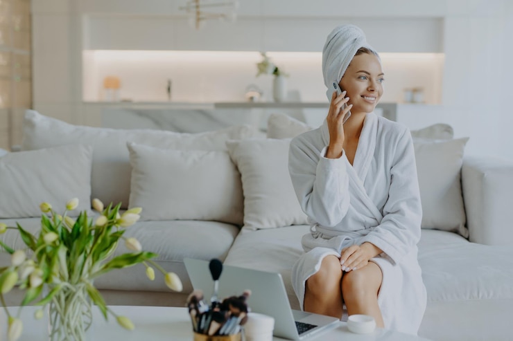 Foto fröhliche schöne frau sieht mit zahnigem lächeln beiseite, hat telefongespräche im bademantel und eingewickeltes handtuch auf dem kopf, umgeben von modernen geräten, verwendet kosmetische produkte, um sich um sich selbst zu kümmern