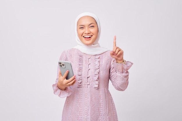 Fröhliche schöne junge asiatische Muslimin mit Hijab und lila Kleid mit Smartphone, die eine Idee hat und nach oben zeigt, isoliert auf weißem Studiohintergrund