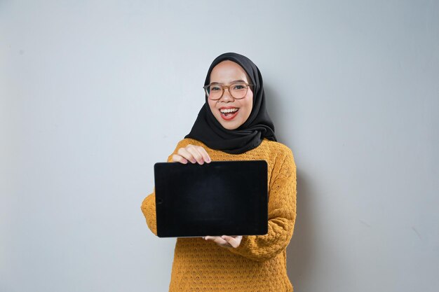 Fröhliche, schöne junge asiatische Muslimin mit orangefarbenem Pullover und Brille, während sie ein Tablet benutzt