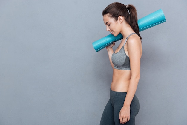 Fröhliche schöne junge Fitnessfrau, die Yogamatte hält und auf grauem Hintergrund wegschaut