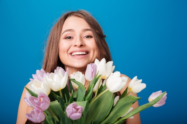 Fröhliche schöne junge Frau, die einen Strauß Frühlingsblumen über blauem Hintergrund lächelt und hält