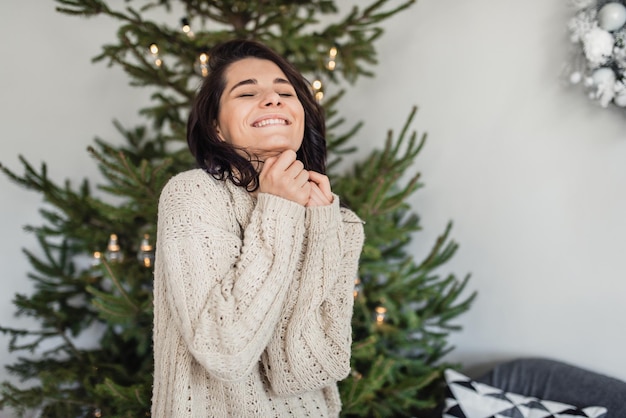 Fröhliche, schöne junge Frau, die neben dem Weihnachtsbaum lächelt und mit geschlossenen Augen auf eine Geschenküberraschung wartet. Kaukasische Frauen, die den Weihnachtsmorgen feiern, posieren gegen Weihnachtsdekoration