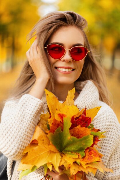 Fröhliche, schöne junge Frau in einem modischen Vintage-Strickpullover und einer Sonnenbrille hält einen schönen hellen Herbststrauß aus Ahornblättern und geht im Freien spazieren