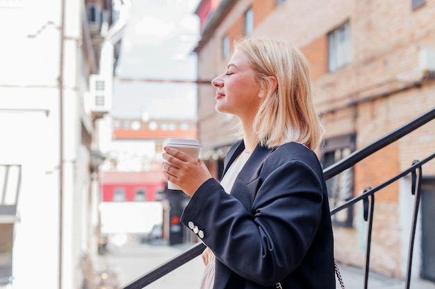 Fröhliche, schöne junge Frau in Kleid und Jacke geht durch die Stadt und trinkt Kaffee