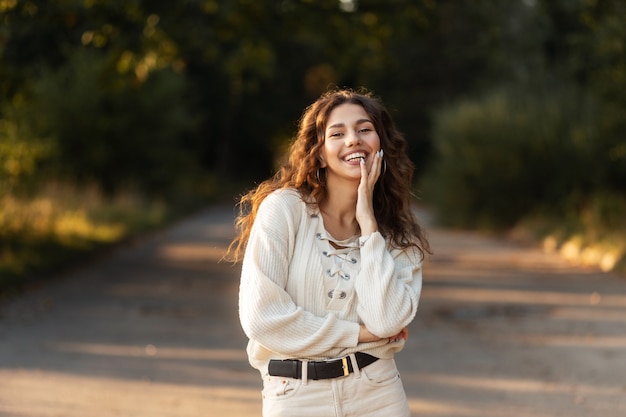 Fröhliche schöne junge Frau mit lockigem Haar und einem schönen Lächeln mit Zähnen in einem Strickpullover im Park in der Natur. Weibliches hübsches lächelndes Gesicht