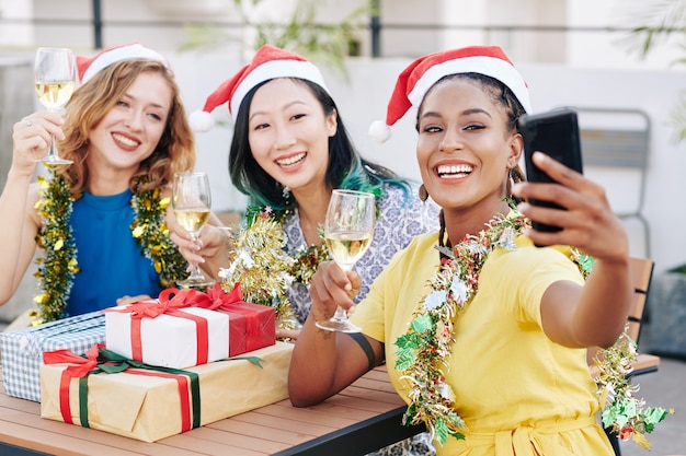 Fröhliche schöne junge hispanische Frauen mit Weihnachtsmann-Hut machen Selfie mit Freunden auf der Weihnachtsfeier, wenn sie Champagner trinken und Geschenke austauschen
